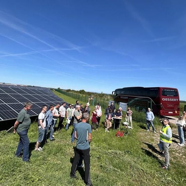 Menschen versammeln sich auf einer Wiese mit Freiflächen-PV und einem Bus im Hintergrund