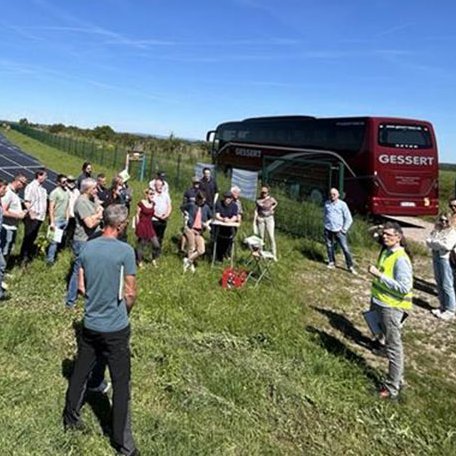 Menschen versammeln sich auf einer Wiese mit Freiflächen-PV und einem Bus im Hintergrund