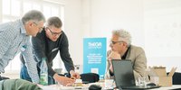 In einem hellen Raum reden drei Personen über ein fachlichen Sachverhalt, zwei Personen stehen und erklären, die dritte Person sitzt und hört zu. 