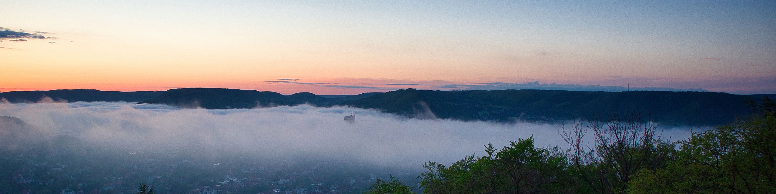 Luftaufnahme von Jena bei Sonnenuntergang mit goldfarbenem Himmel, beleuchteten Gebäuden, der Saale und den umliegenden Hügeln.