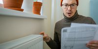 Junger Mann hält schockiert die Nebenkostenabrechnung in der Hand. Die andere Hand liegt auf einem Heizkörper.