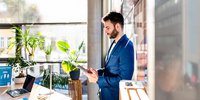 Mann steht in einem Büro vor einem großen Fenster und liest auf einem Tablet die Angebote der Landesenergieagentur ThEGA