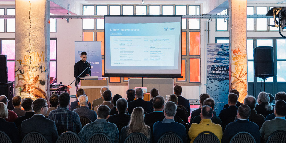 Die Szene wurde von hinten fotografiert, es sitzen ca. 150 Leute im Raum vor einer Leinwand auf der eine Präsentation von Robert Kraft auf dem ThAWI Netzwerktreffen im Kontor Erfurt 2026 läuft. Robert Kraft der ThEGA steht auf der Bühne.