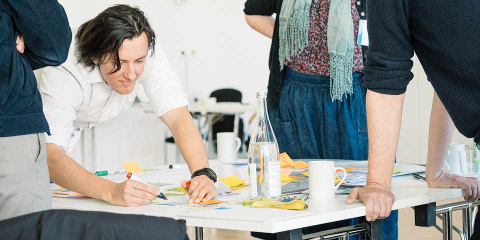 Workshop Gruppenarbeit mit Notizzetteln Ein Gruppe von vier Personen steht um einen weißen Tisch, ein Mann aus der Gruppe schreibt etwas auf einen Notizzettel.